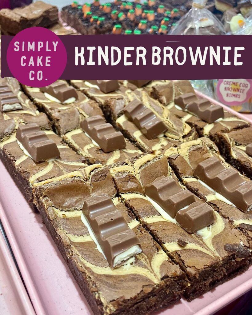 Close-up of Simply Cake Co’s Kinder Brownie slices topped with creamy white chocolate swirls and mini Kinder bars, displayed on a pink tray.