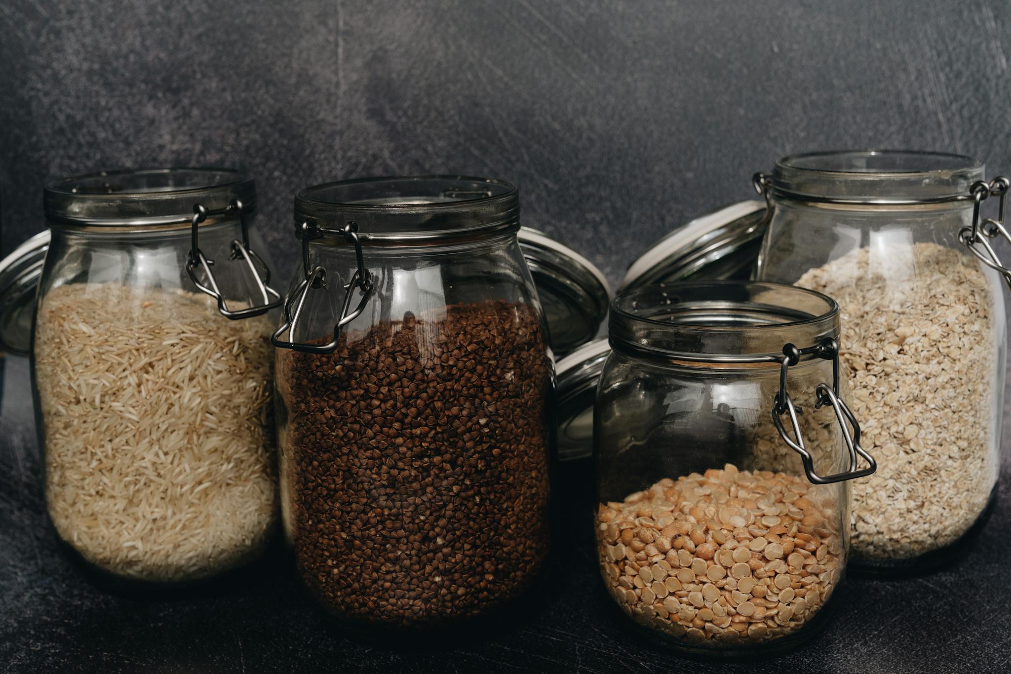 Four glass jars filled with buckwheat, lentils, rice, and oats on a dark surface, ideal for pantry organization.