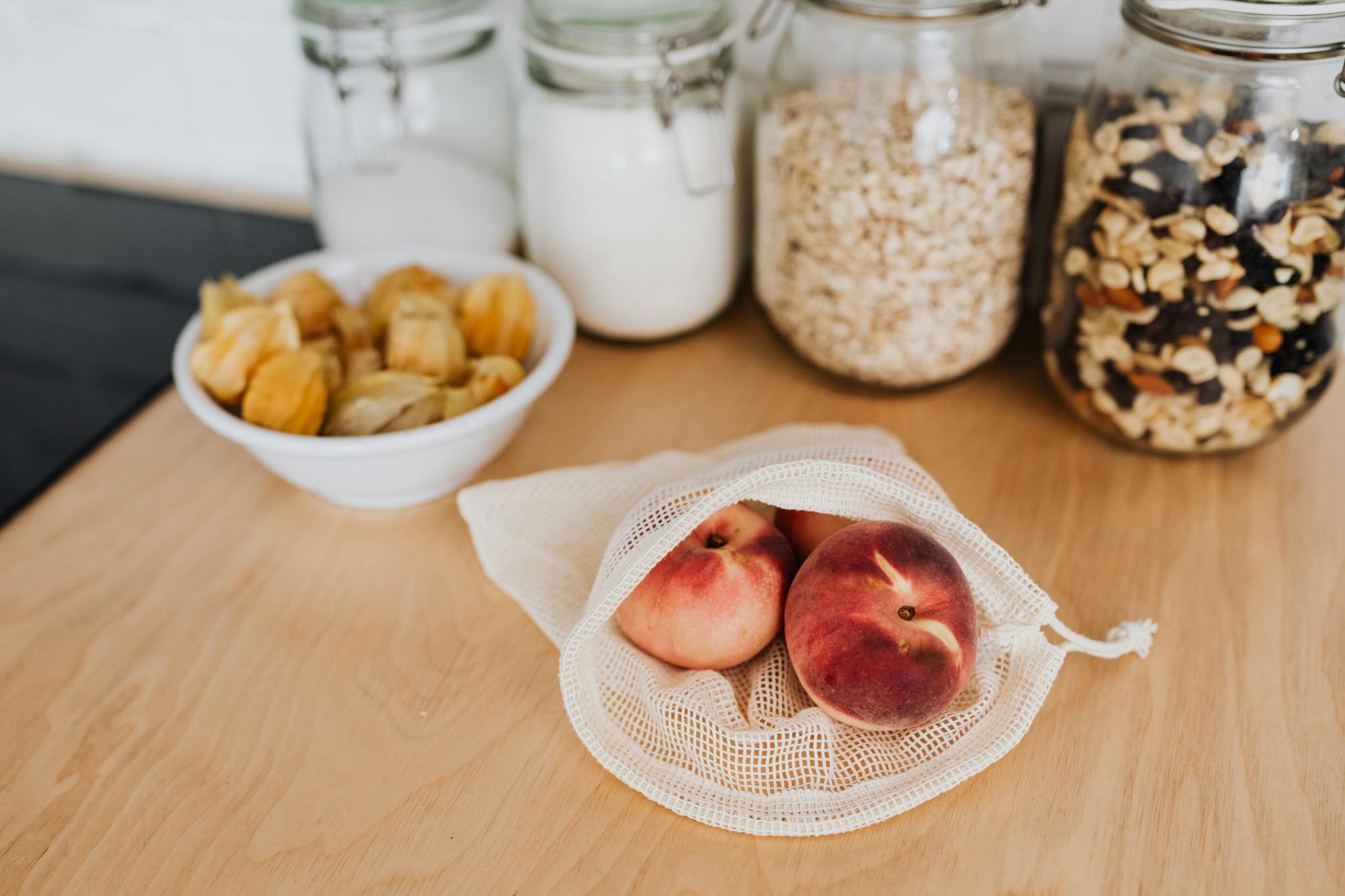 Peaches in a mesh bag on a wooden kitchen countertop with jars of ingredients.