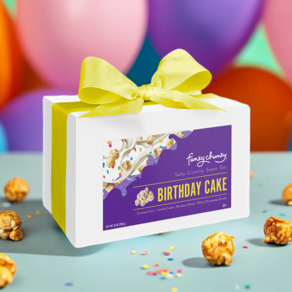Funky Chunky Birthday Cake popcorn gift box with yellow ribbon, featuring caramel corn coated in vanilla cream, rainbow glitter, and white chocolate drizzle, set against a colorful balloon background.